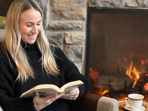 a woman reading a book in front of a fireplace at voco Saranac Lake NY - Waterfront by IHG in Saranac Lake