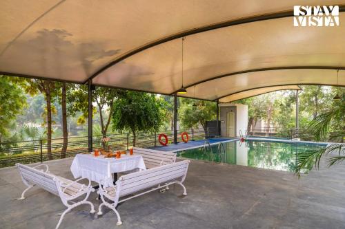 a patio with two white benches and a pool at StayVista at Leela Retreat with BBQ, Bonfire, Pool & Lawn in Sohna