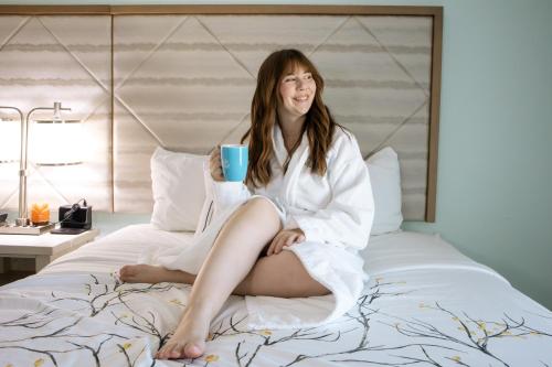a woman sitting on a bed with a cup of coffee at Ocean Sands Beach Boutique Inn-1 Acre Private Beach-St Augustine Historic-2 Miles-Shuttle with Downtown Tour-HEATED Salt Water Pool until 420AM-Popcorn-Cookies-New 4k USD Black Beds- 69 Item Breakfast-Eggs-Bacon-Starbucks- Free Laundry- #904-799-SAND in Saint Augustine