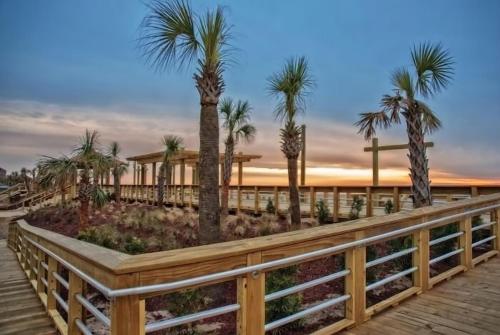a wooden boardwalk with palm trees and a building at Next Time Down by Carolina Beach Realty in Kure Beach