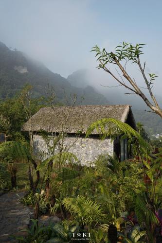 a stone building with a straw roof with mountains in the background at TOKI Ecolodge Cao Răm in Hòa Bình