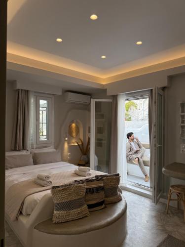 a man sitting in a chair in a bedroom looking out the window at Fratelli Family Rooms in Tinos Town