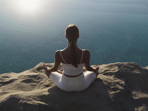 a woman sitting in meditation on a rock near the water at Magma Resort Santorini, In The Unbound Collection By Hyatt in Vourvoulos