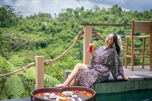 Una donna seduta sul bordo di una piscina con un drink in mano di The Scape Valley Villas by GenuineHost a Tampaksiring