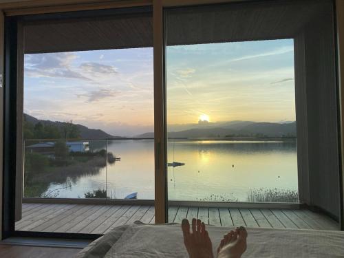 a person laying on a bed looking out a window at a lake at Villa Über-See with private beach in Pörtschach am Wörthersee