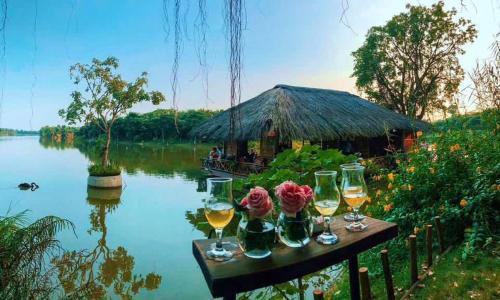 a table with glasses of wine and a hut on a river at Vườn Tùng RiverSide retreat & Villa - KDT ECOPARK in Kim Quan