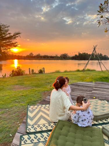 a woman and two children sitting on a blanket next to a lake at VAA chachoengsao in Ban Hua Sai