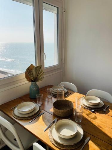 une table en bois avec des assiettes et des chaises et une fenêtre dans l'établissement L'Escapade, à Fort-Mahon-Plage