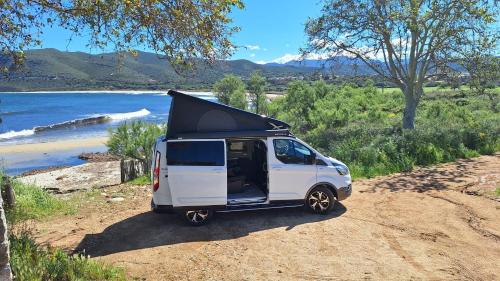 une camionnette blanche garée sur un chemin de terre avec sa porte ouverte dans l'établissement Van&Scoot, à Belgodère