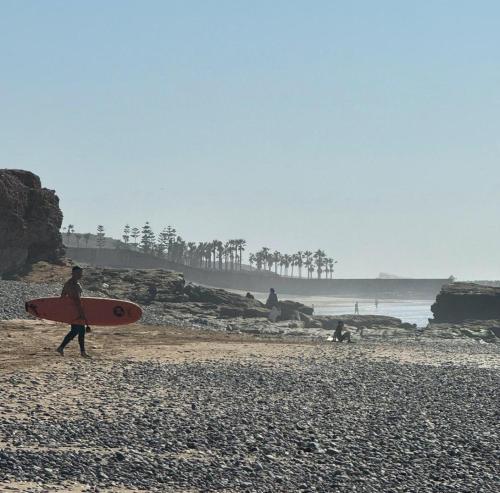 a man walking on the beach with a surfboard at Tasga Guest House in Tamraght Oufella