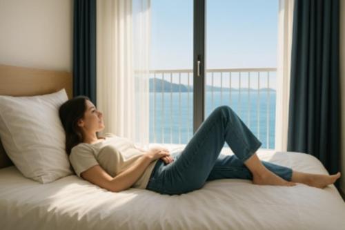 a woman sitting on a bed looking out a window at Yeosu U-Bless Terrace Ocean View in Yeosu