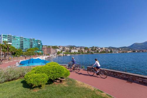 two people riding bikes on a sidewalk next to a body of water at Viganello Modern Apartments in Lugano