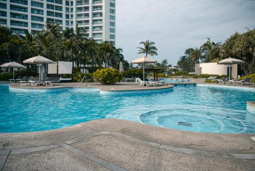a large swimming pool with chairs and trees and buildings at Departamento Mayan Lakes T9 - Albercas, club de playa, tenis, pádel y más in Acapulco