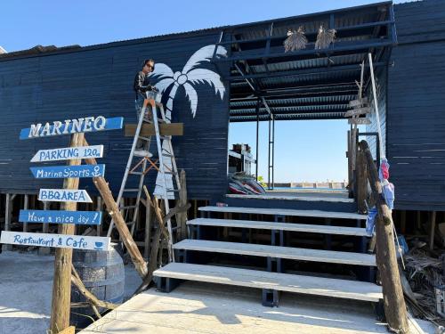 a man on a ladder painting a blue building at MARINE-Q in Inami