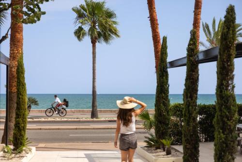 a woman wearing a hat standing on a sidewalk looking at the ocean at Hotel Royalmar - Nouveau propriétaire in Cagnes-sur-Mer
