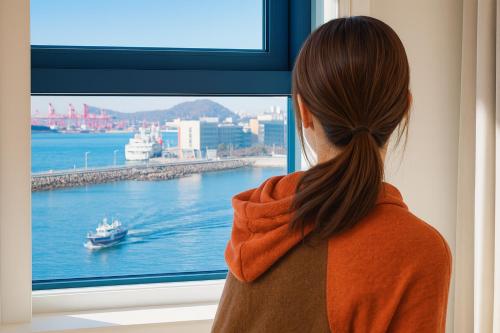 a woman looking out a window at a boat in the water at EL Stay Hotel Residence in Busan