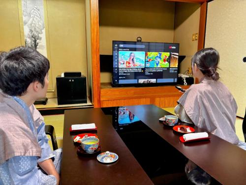 a man and a woman sitting at a table watching a television at Yufuin Santoukan in Yufuin