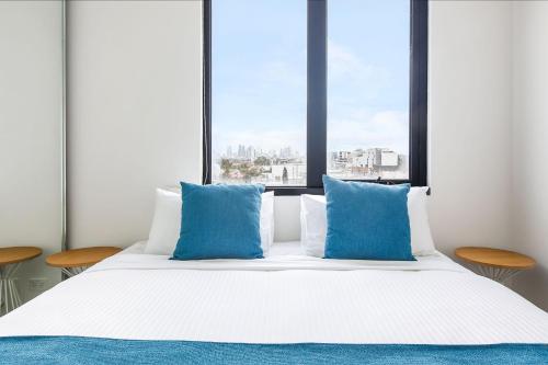 a white bed with blue pillows in front of a window at Artel Apartments Brunswick in Melbourne