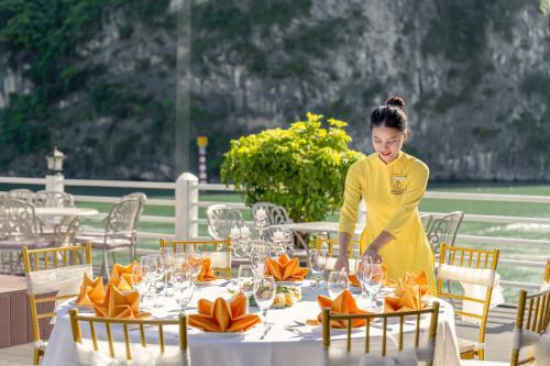 a woman in yellow is setting a table with glasses at Athena Royal Cruise - Luxury in Halong Bay in Ha Long
