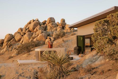a person standing next to a house in front of a mountain at Fox Inn Epic Views, Hot Tub, Cowboy Tub, Grill in Yucca Valley