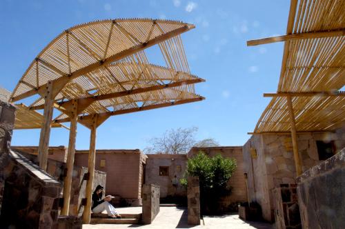 a woman sitting under a wooden structure in front of buildings at Hotel Kimal in San Pedro de Atacama