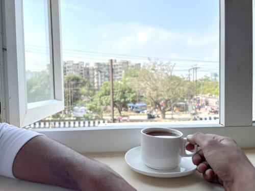 a person holding a cup of coffee in front of a window at Hotel Mehak Palace By Aura in Noida