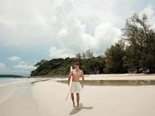 a man standing on a beach holding a surfboard at The Survival by Seudam in Ko Kood