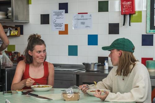 two women sitting at a table in a kitchen eating food at Sagres Sun Stay - Surf Camp & Hostel in Sagres