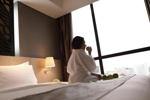 a woman sitting on a bed drinking from a cup at Hotel Grandhika Pemuda in Semarang