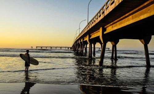 a man standing on the beach with a surfboard under a pier at Casa Com Piscina - Mongaguá - Disponível Feriado Carnaval in Mongaguá