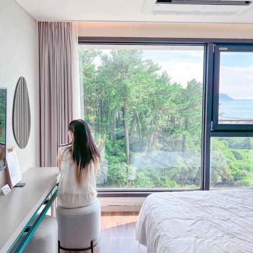 a woman sitting at a desk in a bedroom looking out a window at Hotel Seogwipean in Seogwipo