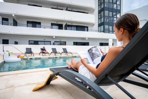 a woman sitting in a chair reading a book by a pool at Nemea Appart Hotel Les Hauts de Milady Biarritz in Biarritz