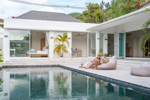 two women sitting next to a swimming pool in front of a house at Enchanting 4Br Villa Private Pool & Breakfast incl in Mae Nam