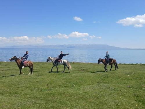 three people riding horses on the grass near the water at Jusup Guest House, Hostel in Dzhangyaryk