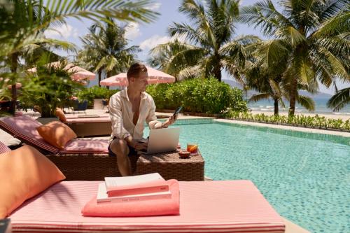 a man sitting on a chair by a pool with a laptop at Signature by M Village Beachfront Hotel in Da Nang