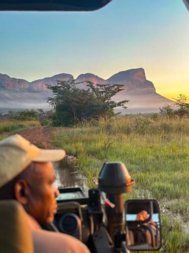 two people in a jeep on a road in a field with mountains at Honeyguide Conservation Lodge in Golders Green