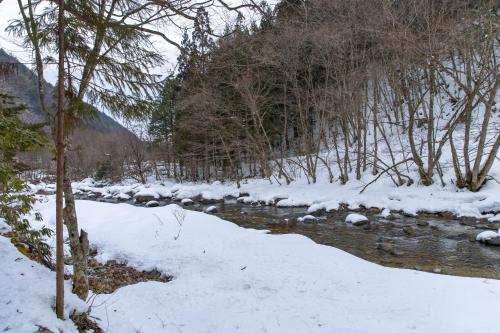 a stream with snow on the ground and trees at Wanosato in Takayama