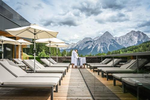 a man standing on a deck with lounge chairs and mountains at Hotel Lärchenhof in Lermoos