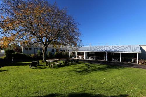 a building with picnic tables and a tree in a field at Waiuku Lodge Motel in Waiuku