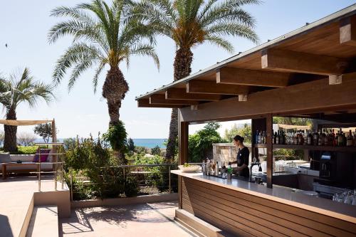 a woman sitting at a bar on a patio with palm trees at Aquagrand Artistic Luxury Beach Resort - Adults Only in Líndos