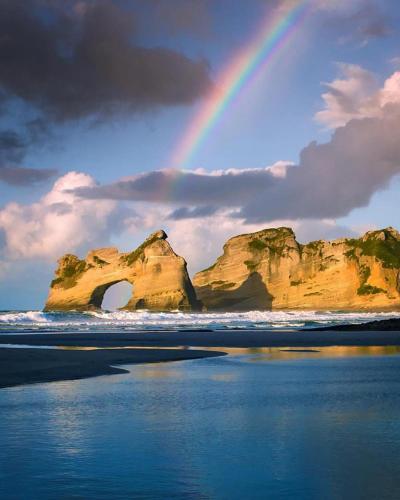 a rainbow over a beach with a rock arch in the water at Annies Nirvana Lodge Backpackers in Takaka