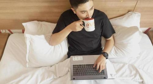 a man sitting in bed with a cup of coffee and a laptop at Evora Hotel in Surabaya