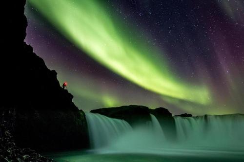 a person standing next to a waterfall under the northern lights at LUXURIOUS COMFORTABLE Family Villaes in Akureyri