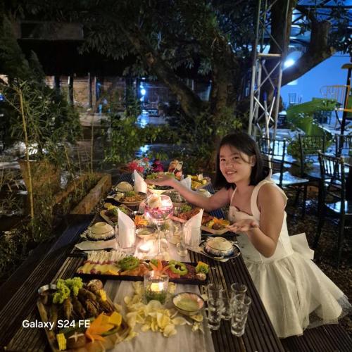 a woman in a white dress sitting at a table with food at Homestay Thác Liên Khương in Ðưc Trọng