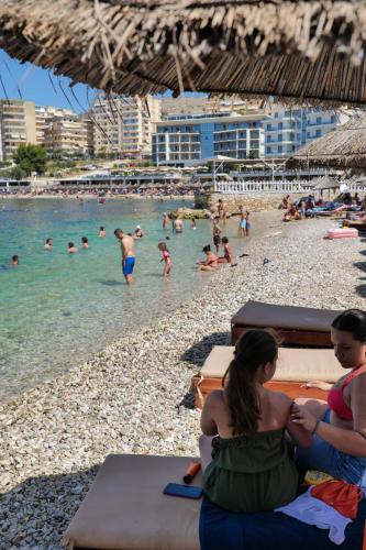 a woman sitting on a beach with people in the water at Sunset Aparthotel in Sarandë