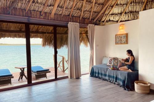 a woman sitting on a bed in a room with the ocean at Akalki Hotel y Centro Holistico in Bacalar