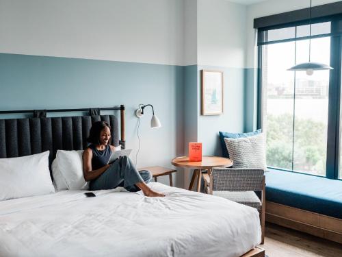 a woman sitting on a bed with a laptop at The Junto Hotel in Columbus