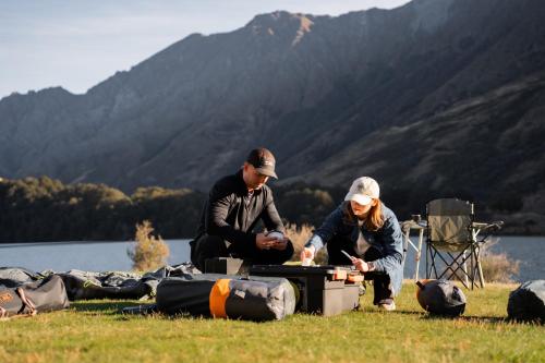 two people sitting on the grass near a lake at Rent-A-Tent - Hire camping gear in Auckland