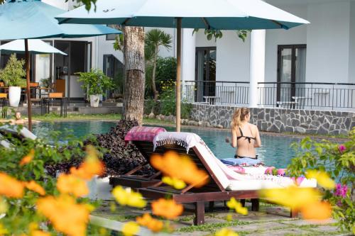 a woman in a bikini sitting on a chaise lounge by a pool at ÊMM Hotel Hoi An in Hoi An