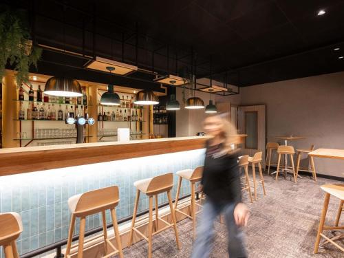 a woman walking past a bar in a restaurant at Novotel Macon Nord - Porte de Bourgogne in Mâcon